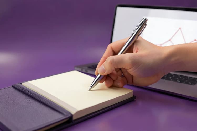 Uma mão escreve em um caderno fechado ao lado de um laptop com um gráfico simples na tela.
