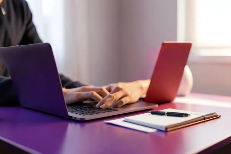 Uma pessoa trabalhando em um laptop em uma mesa com um caderno e uma caneta ao lado